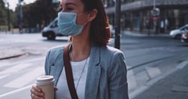 Koruyucu maskeli bir kadın kahve içiyor, şehirde yürüyor. Yavaş bir hareket yap. Genç bayan hava kirliliğine ve Coronavirus covid-19 'a karşı maskesini çıkararak kağıt bardaktan su içiyor..