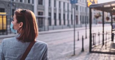 Koruyucu Maskeli Genç Kadın Güneşli Sabah Şehrinde Tramvay Bekliyor. Yavaş hareket, Gimbal Dengeleyici. Hava kirliliği ve koronavirüse karşı maskeli iş kadını toplu taşıma kullanıyor.