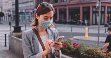 Şehirde koruyucu maske takan, akıllı telefon kullanan bir kadın var. Yavaş hareket, Gimbal Dengeleyici. Hava kirliliği ve Coronavirus Covid-19 'a karşı maskeli genç bir kadın. Şehir Sabahı.