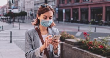 Şehirde koruyucu maske takan, akıllı telefon kullanan bir kadın var. Yavaş hareket, Gimbal Dengeleyici. Hava kirliliği ve Coronavirus Covid-19 'a karşı maskeli genç bir kadın. Şehir Sabahı.
