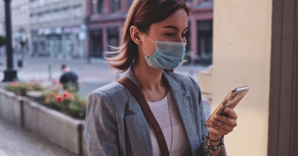Woman Wearing A Protective Mask Walking In The City, Using Smartphone. SLOW MOTION, Gimbal Stabilizer. Young Female in face mask against air pollution and coronavirus Covid-19. Urban Morning. — Stock Video