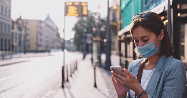 Junge Frau in Schutzmaske mit Smartphone, die in der sonnigen Morgenstadt auf die Straßenbahn wartet. SLOW MOTION, Gimbal Stabilisator. Geschäftsfrau mit Mundschutz gegen Luftverschmutzung und Coronavirus. — Stockvideo
