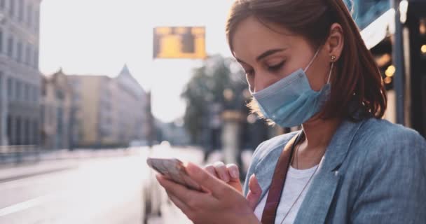 Young Woman In Protective Mask Using SmartPhone, Waiting For Tram In Sunny Morning City. SLOW MOTION, Gimbal Stabilizer. BusinessWoman in face mask against air pollution and coronavirus use transport. — Stock Video