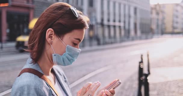 Femme portant un masque protecteur marchant dans la ville, utilisant un téléphone intelligent. Lentement, stabilisateur de cardan. Jeune femme masquée contre la pollution de l'air et le coronavirus Covid-19. Matin urbain. — Video