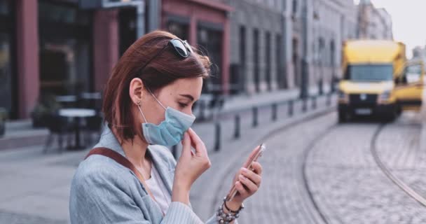 Femme portant un masque protecteur marchant dans la ville, utilisant un téléphone intelligent. Lentement, stabilisateur de cardan. Jeune femme masquée contre la pollution de l'air et le coronavirus Covid-19. Matin urbain. — Video