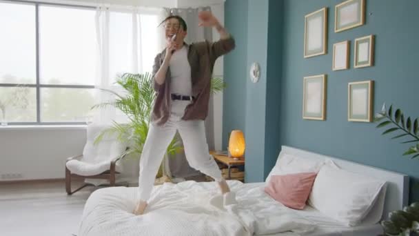 幸せな女性は家でベッドの上でジャンプ踊り 面白いダンスでお祝いを楽しんでいます 電話でカラオケを歌い ベッドの上に落ちる — ストック動画