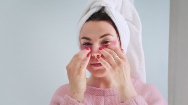 Aynanın önünde duran bir kadın gözlerinin altına hidrojel kolajen pembe bantlar yapıştırıyor. Nemlendirici ve yaşlanmayı önleyici cilt bakımı. Koyu halkalar için pedler