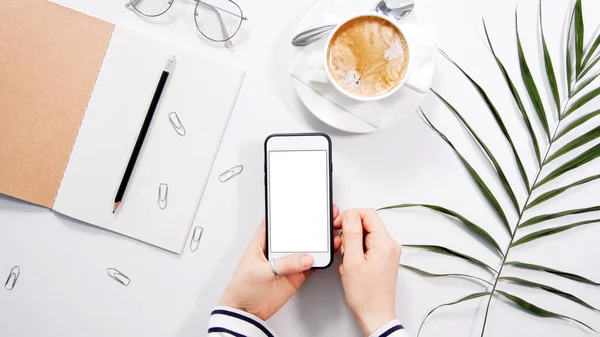 Flatlay on white work space desk with woman\'s hands holding white smartphone with white copyspace, coffee, planner and other office accessories. Mobile phone mockup.