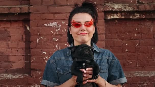 身穿时髦红色眼镜的时髦女人笑着在怀里摇动一只滑稽的哈巴狗 肖像学生 千禧年爱犬 — 图库视频影像