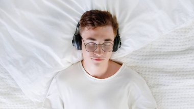Sakin genç adam, evde stres hissetmeden meditasyon yaparken dinlenmeyi seviyor. Öğrenci erkek, yatakta gözleri kapalı ve kulaklık takıyor.. 