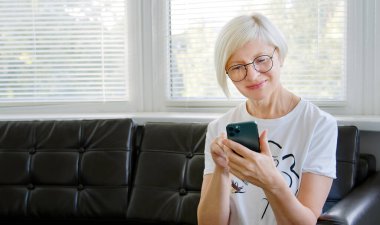 Telefonu kullanan olgun bir kadın. Orta yaşlı bir kadın elinde akıllı bir telefonla cep telefonu ekranına bakıyor sosyal medya uygulamaları kullanarak mobil oyunlar oynuyor ve evdeki koltukta oturuyor. Yaşlılar ve modern teknoloji kavramı.