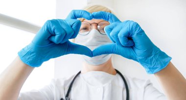 Tıbbi ceketli, maskeli ve steteskoplu gülümseyen kadın doktor ellerin kalp şeklini gösteriyor. Tıp, aşk işi, sağlık hizmeti konsepti. Seçici odak.