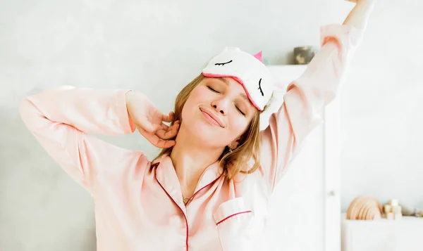 Pembe pijamalı ve uyku maskeli genç bir kadın, uyanıp uykusundan sonra geriniyor, uykulu ve mutlu. İyi ve sağlıklı uyku konsepti.