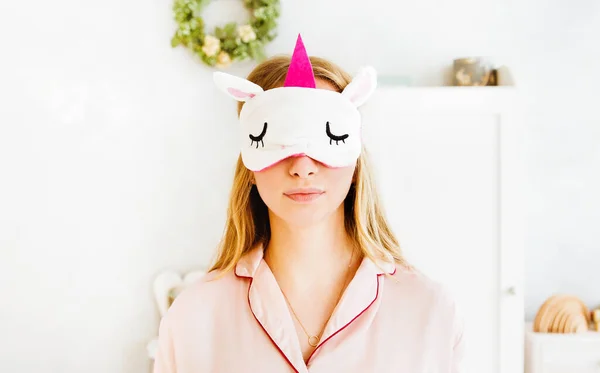 Uyku maskeli tek boynuzlu at ve pembe pijama giymiş yatak odasında poz veren genç bir kadın.. 