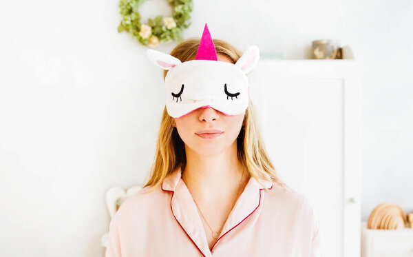 Young woman in sleep mask unicorn and pink pajama posing on bedroom background. 
