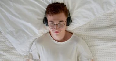 Sakin genç adam, evde stres hissetmeden meditasyon yaparken dinlenmeyi seviyor. Öğrenci erkek, yatakta gözleri kapalı ve kulaklık takıyor.. 