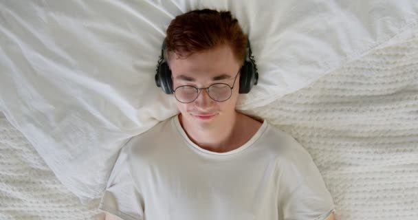 落ち着いた若者は 冷たい音楽のオーディオサウンドを聞いて自宅でストレスを感じる瞑想を楽しんでいます 学生男性リラックスベッドで目を閉じて ヘッドフォンを身に着けて — ストック動画