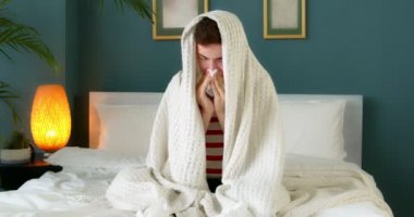 Genç hasta bir adam mendilin içinde hapşırıyor. Burnunu sümkürecek. Üstü örtülü bir şekilde evde oturuyor. Hasta erkek grip oldu ve grip oldu. Alerji belirtileri gösteriyor. Mevsimsel virüs hastalığı.