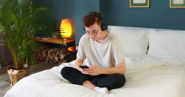 젊은이는 자기가 음악을 침대에 전화와 헤드폰을 사용하여 듣는다 학생들은 스마트폰에서 — 비디오