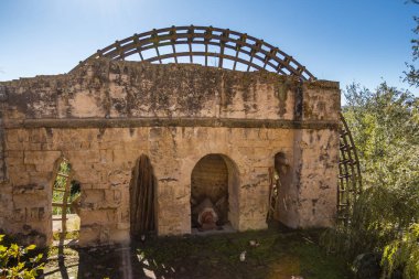 Ancient stone mill with wooden wheel in overgrown field in sunlight in Cordoba, Spain
