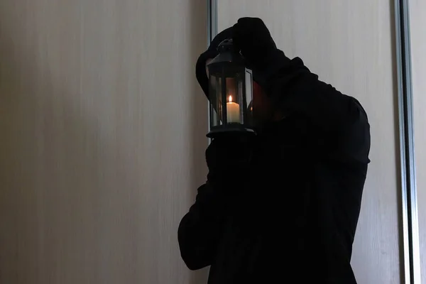 A thief in a black hood and gloves with a lamp with candles in someone else\'s dark apartment. Copy space - the concept of crime, theft, security, protection.