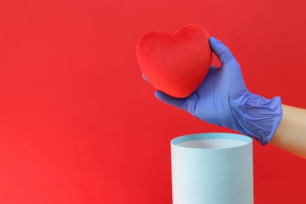 Soft, heart in a hand in a medical glove near a round, gift box on a red background. Copy space - 2019-nCoV pandemic concept, coronavirus, treatment, quarantine, safety, health, care.