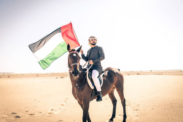 Middle eastern handsome man with typical emirates dress riding a arabic horse in the Dubai desert