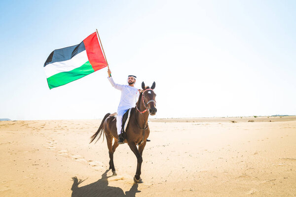 Middle eastern handsome man with typical emirates dress riding a arabic horse in the Dubai desert