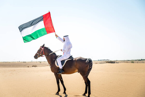 Middle eastern handsome man with typical emirates dress riding a arabic horse in the Dubai desert