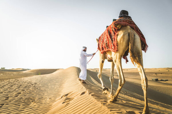 Handsome middle eastern man with kandura and gatra riding on a camel in the desert