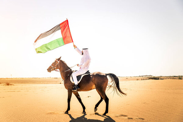 Middle eastern handsome man with typical emirates dress riding a arabic horse in the Dubai desert