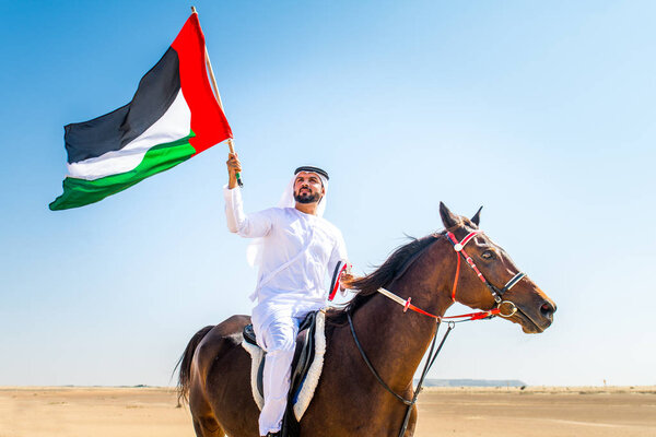 Middle eastern handsome man with typical emirates dress riding a arabic horse in the Dubai desert