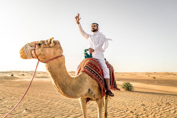 Handsome middle eastern man with kandura and gatra riding on a camel in the desert