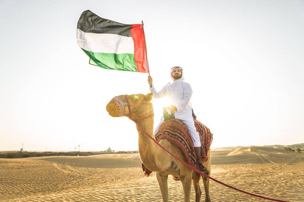 Handsome middle eastern man with kandura and gatra riding on a camel in the desert
