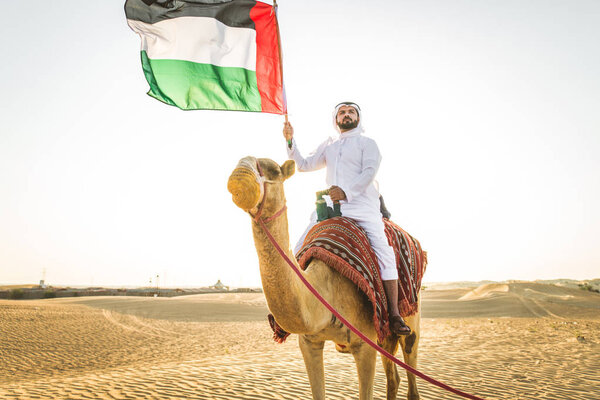Handsome middle eastern man with kandura and gatra riding on a camel in the desert