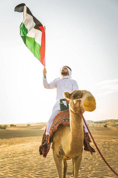 Handsome middle eastern man with kandura and gatra riding on a camel in the desert