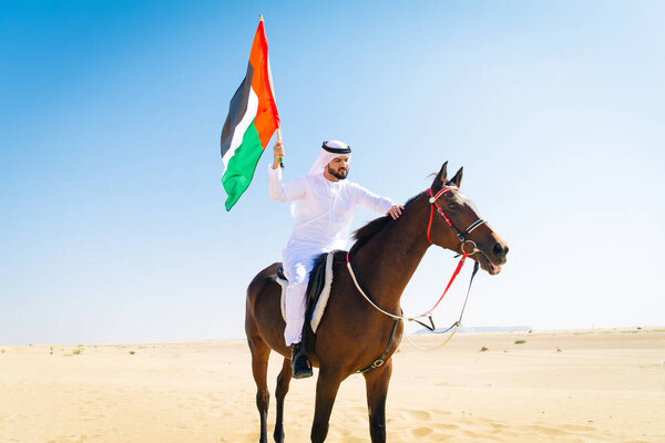 Middle eastern handsome man with typical emirates dress riding a arabic horse in the Dubai desert
