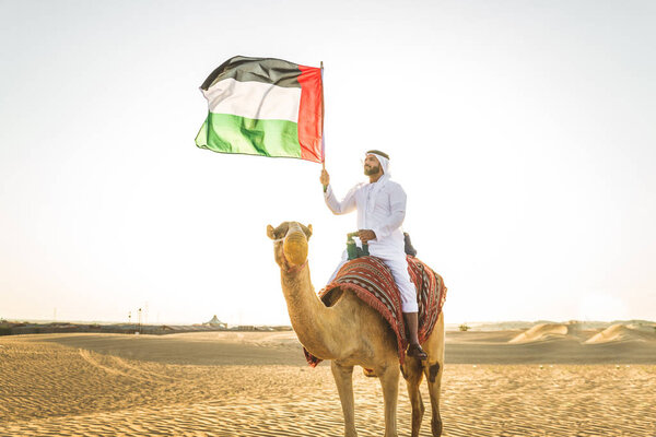 Handsome middle eastern man with kandura and gatra riding on a camel in the desert