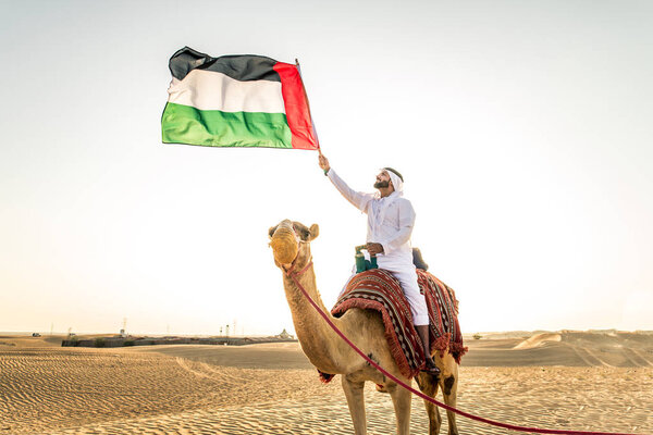 Handsome middle eastern man with kandura and gatra riding on a camel in the desert