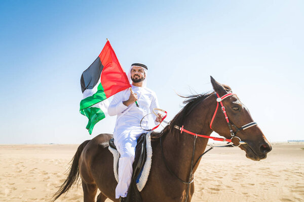 Middle eastern handsome man with typical emirates dress riding a arabic horse in the Dubai desert