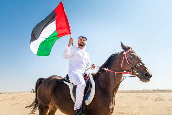 Middle eastern handsome man with typical emirates dress riding a arabic horse in the Dubai desert