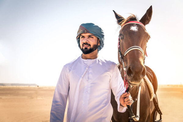 Middle eastern handsome man with typical emirates dress riding a arabic horse in the Dubai desert