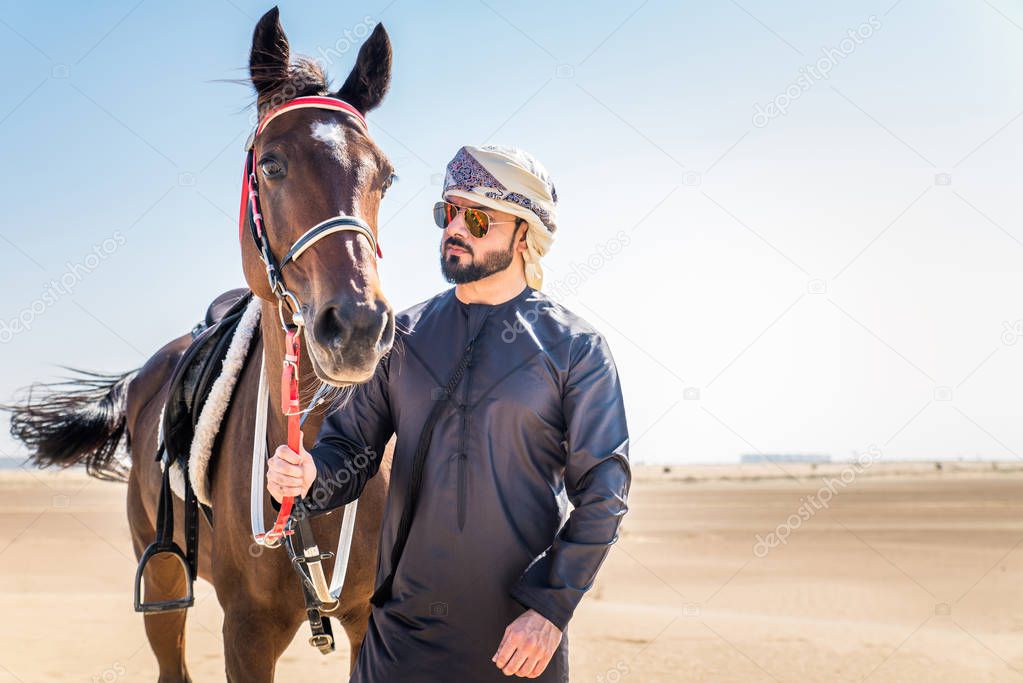 Middle eastern handsome man with typical emirates dress riding a arabic horse in the Dubai desert