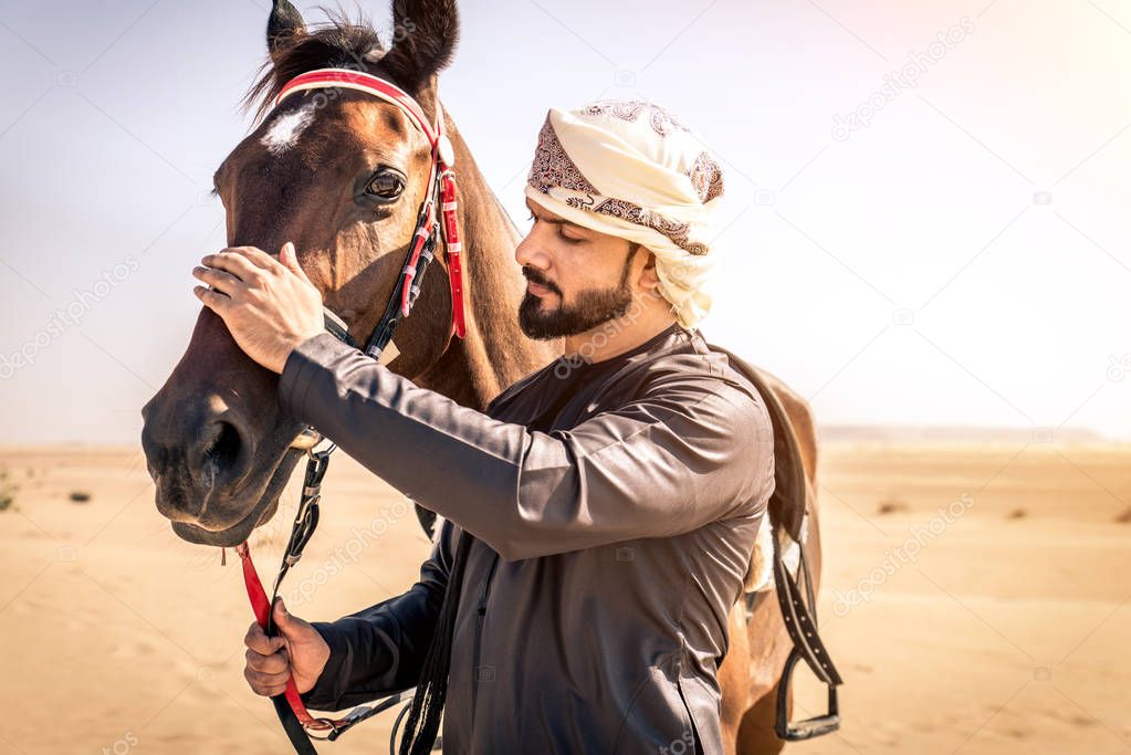 Middle eastern handsome man with typical emirates dress riding a arabic horse in the Dubai desert