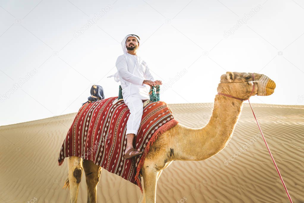 Handsome middle eastern man with kandura and gatra riding on a camel in the desert