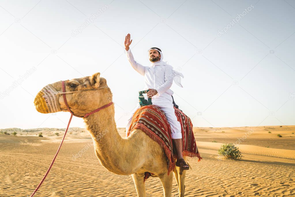Handsome middle eastern man with kandura and gatra riding on a camel in the desert
