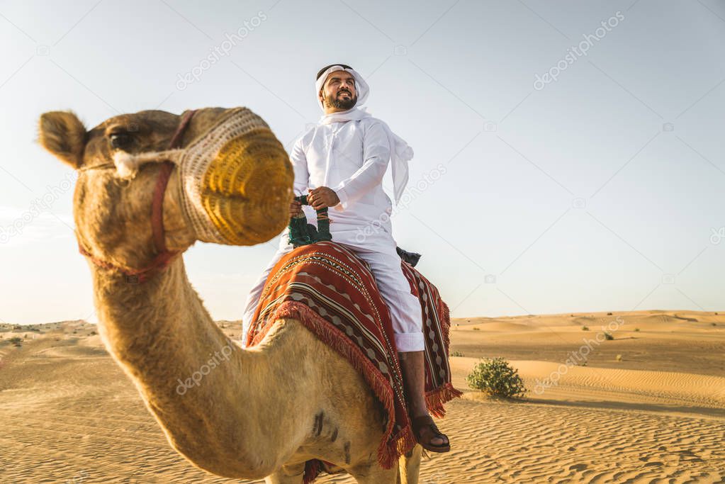 Handsome middle eastern man with kandura and gatra riding on a camel in the desert