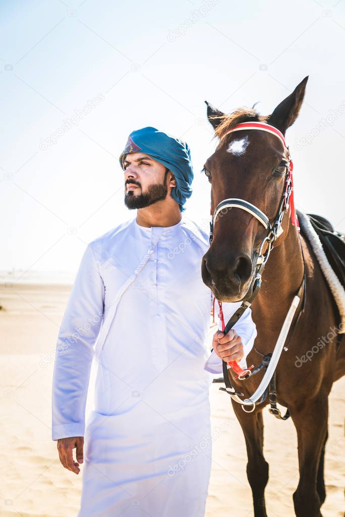 Middle eastern handsome man with typical emirates dress riding a arabic horse in the Dubai desert