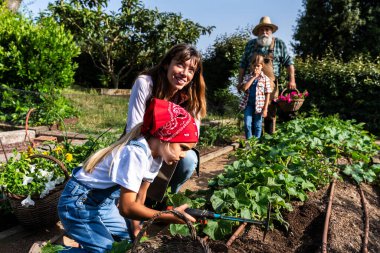 Anne ve kız bitkilerle ilgileniyor, büyükbaba ve diğer kız güneşli bir sebze bahçesinde çiçek sepeti taşıyor, sürdürülebilir tarım ve aile bağlarını destekliyor.