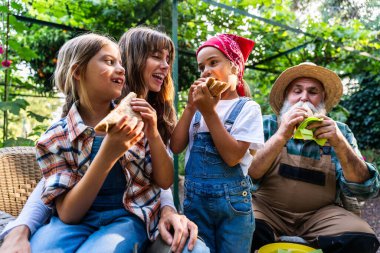 Anne, kızları ve büyükbabasıyla birlikte bir biyoloji çiftliğinin serasında sağlıklı bir sandviç molası veriyor, gülümsüyor ve birbirlerine bakıyorlar.
